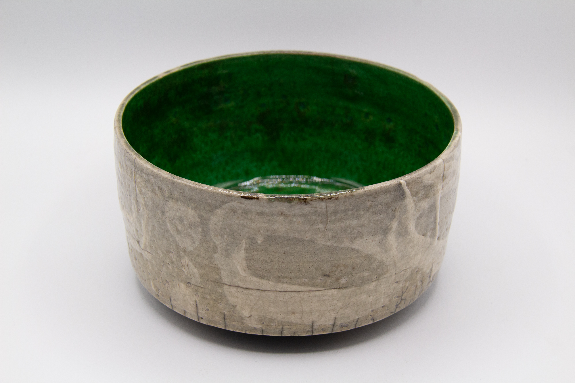 A photograph of a raku fired copper green and clear glazed footed tea bowl with a crackled surface and white slip decoration on the surface by Andrew Cornell Robinson